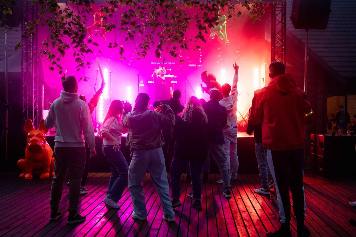 Crowd dances in front of a stage lit by vivid pink and purple lights at a lively outdoor evening party.