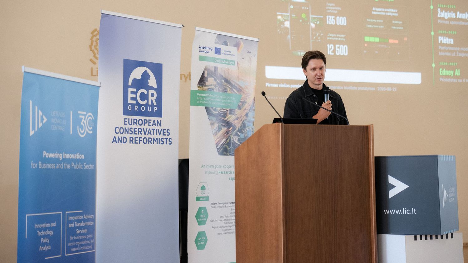 Man speaks into a microphone behind a wooden lectern with ECR Group and Lithuanian Innovation Centre banners at a conference.