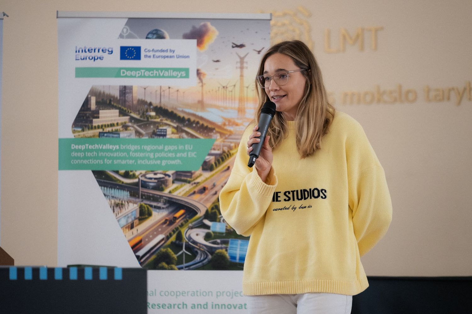 A woman in a yellow sweatshirt speaks into a microphone beside a DeepTechVillage Interreg Europe banner at a conference.