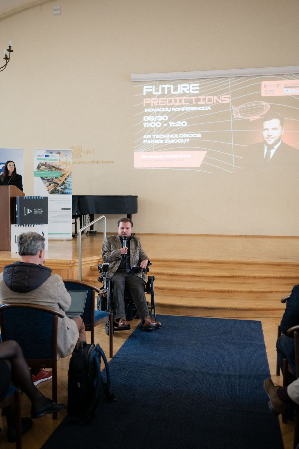 Man in a wheelchair speaks into a microphone on stage as a Future Predictions slide is projected, with attendees watching.