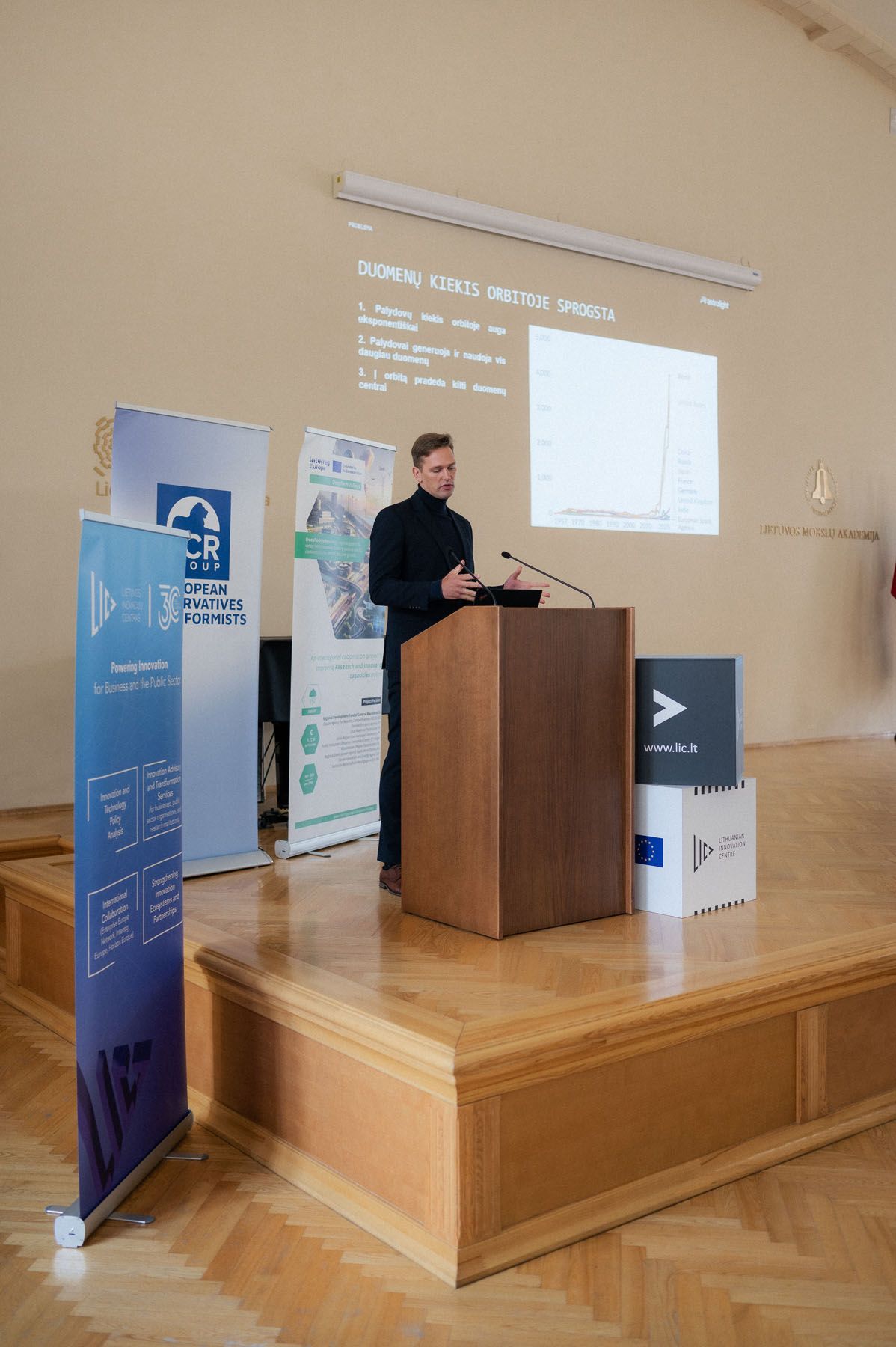 Speaker in dark clothing presents data slides at a wooden podium on stage with European Conservatives banners.