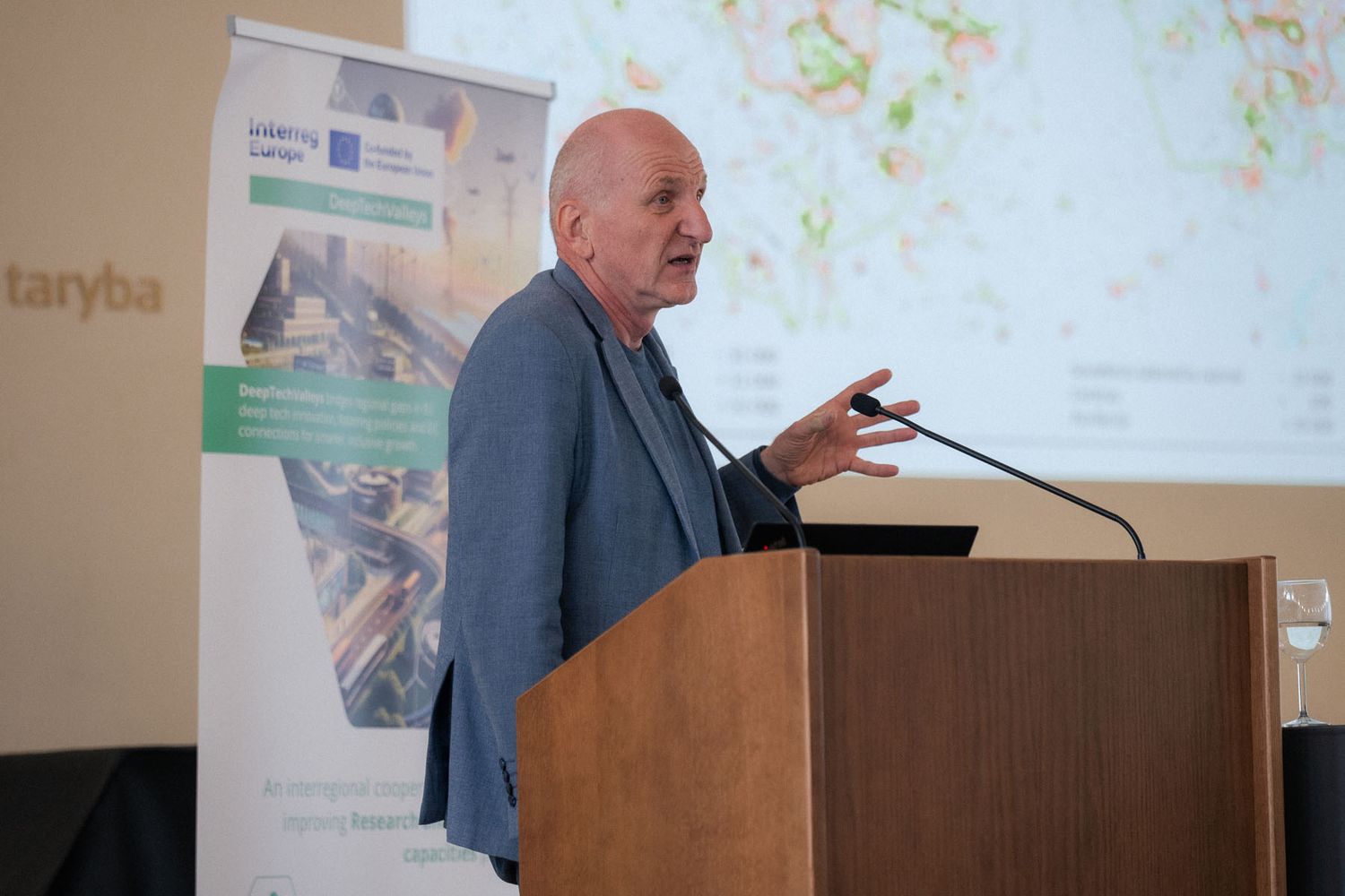 A bald man gestures while speaking into a microphone at a lectern, with a map projected on a screen behind him.