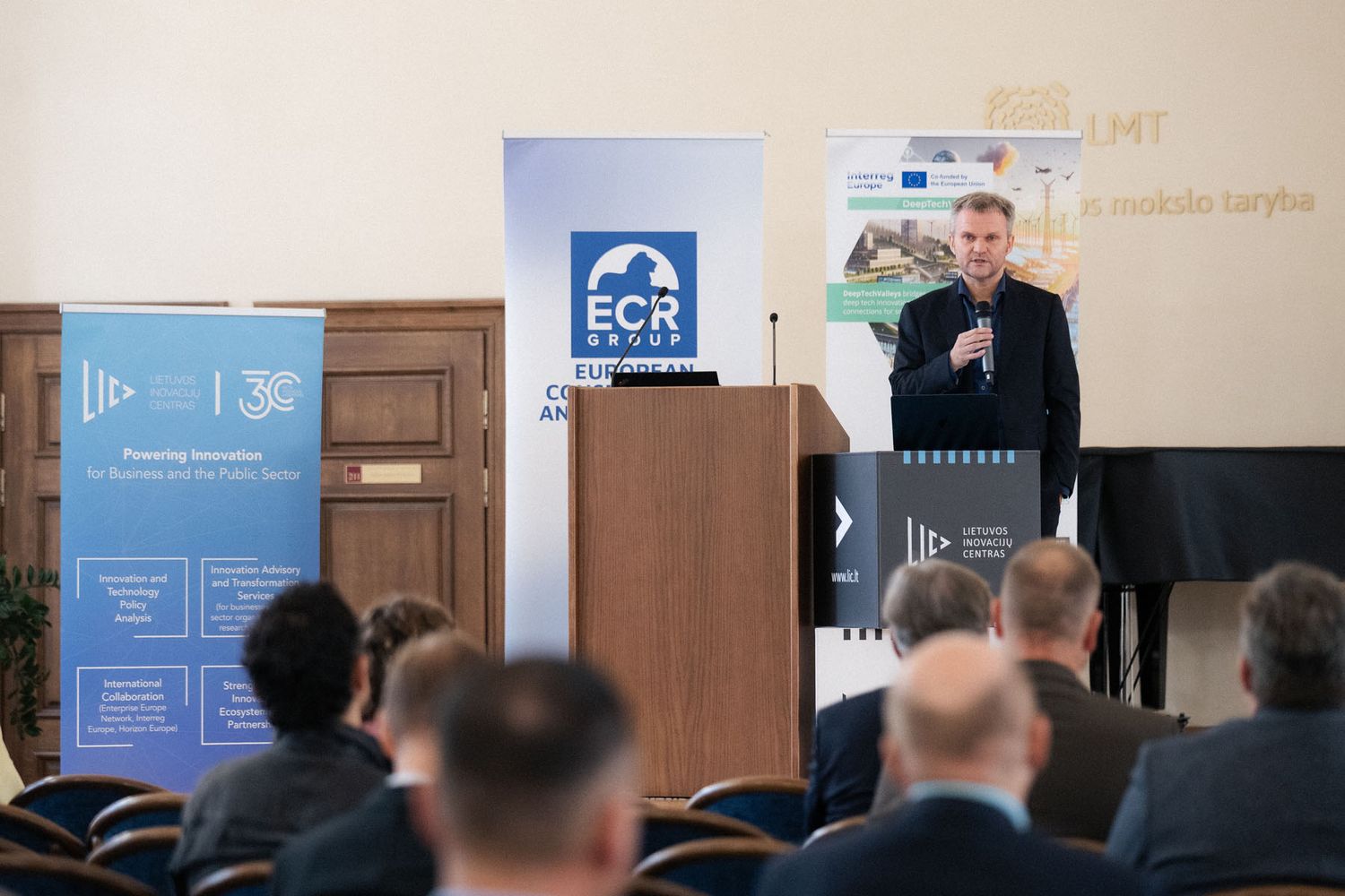 A speaker with a microphone stands near a podium and ECR Group banner while the audience watches from blue chairs.