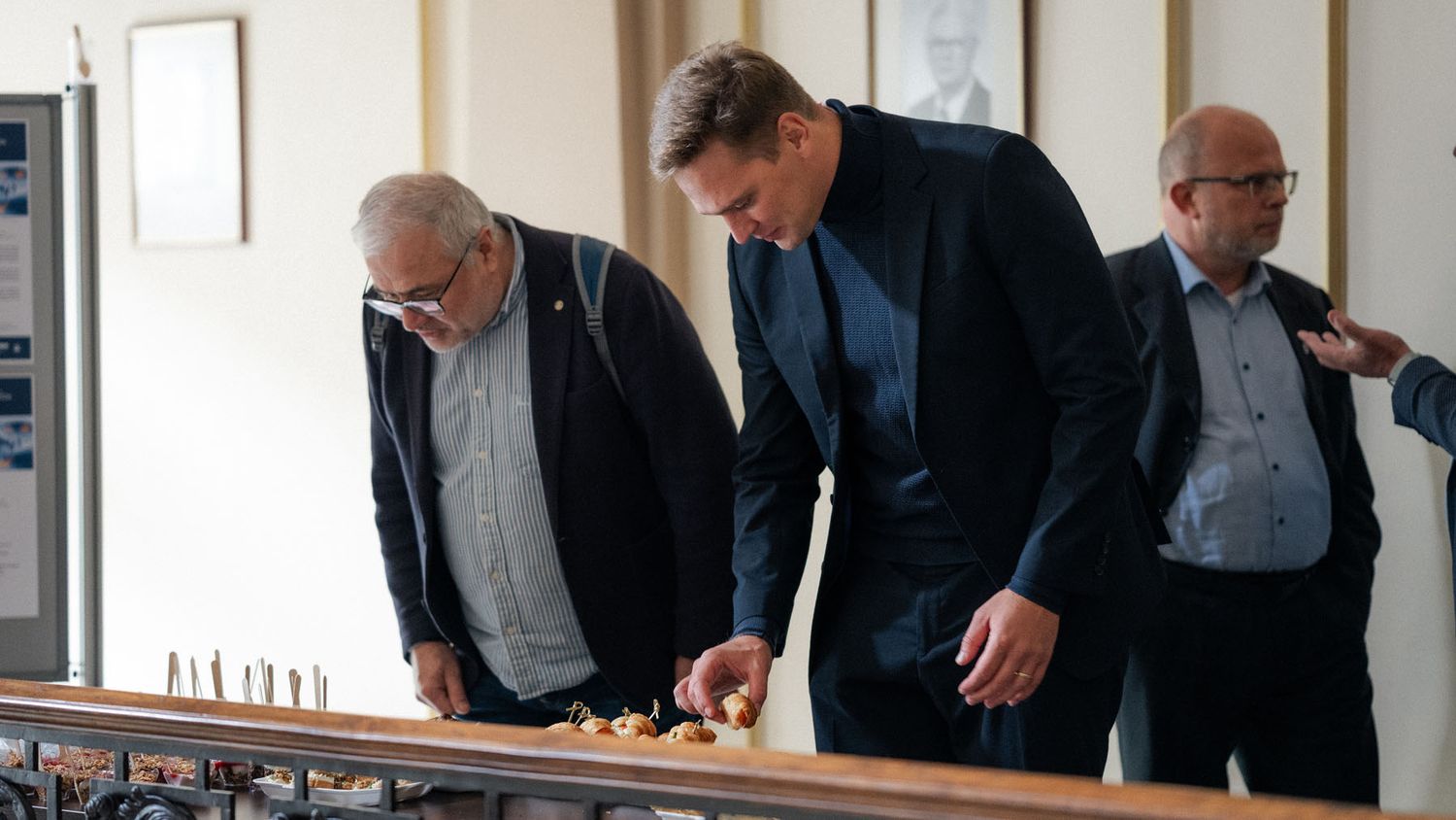 Three men in dark jackets look at items on a wooden railing display during a conference break.