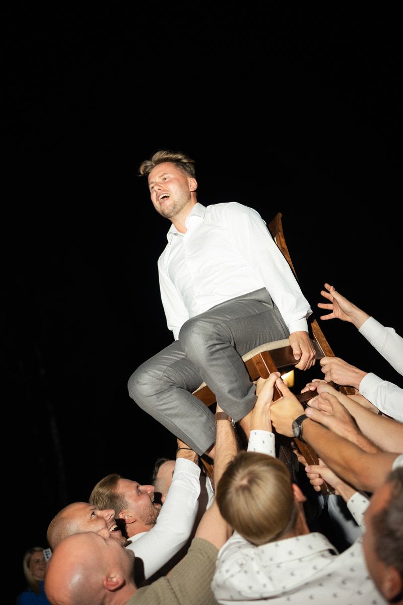 Groom in a white shirt and gray trousers is lifted on a chair by cheering guests during a wedding celebration.