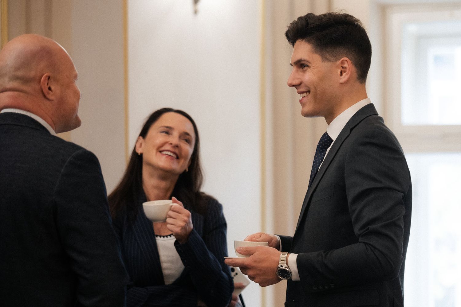Two men in suits and a smiling woman in a dark blazer chat cheerfully while holding cups at a conference networking break.