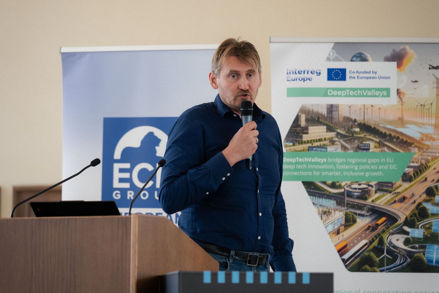 A man in a dark blue shirt speaks into a microphone at a conference with ECR Group and Interreg Europe banners behind him.