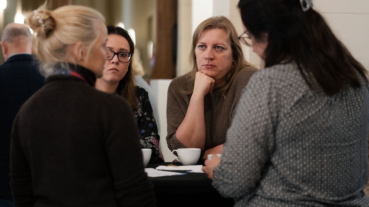 Four women sit around a table with coffee cups, engaged in a thoughtful conversation during a conference break.