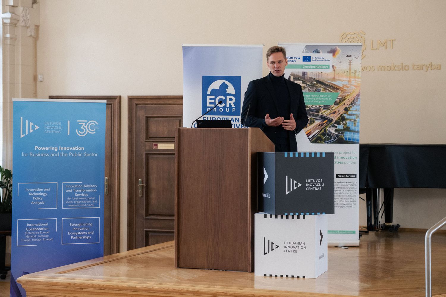 A man in dark clothing speaks at a wooden podium beside Lithuanian Innovation Centre boxes and an ECR Group banner.