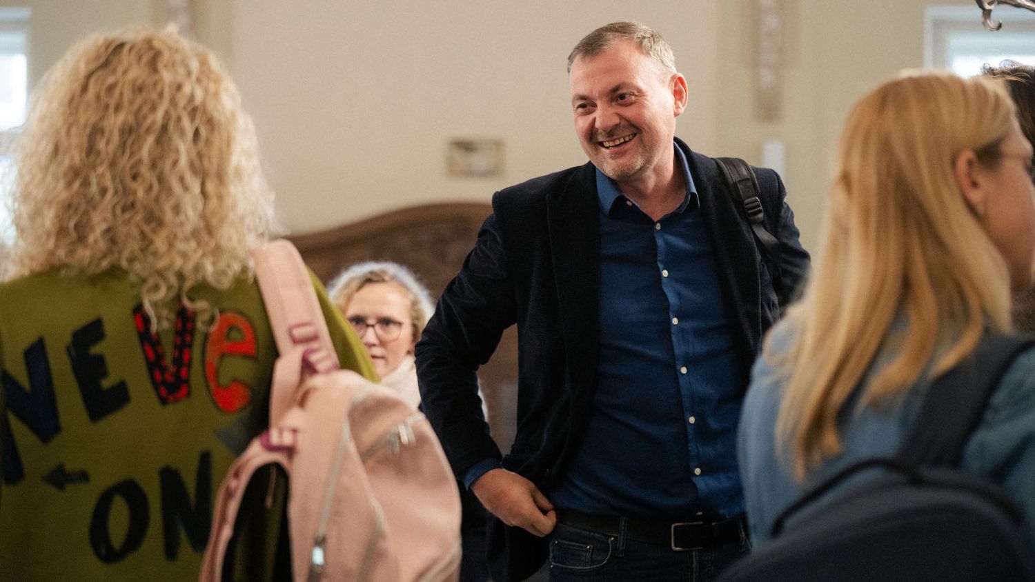 A smiling man in a dark jacket and blue shirt greets conference guests, including a woman in a green sweatshirt with a pink backpack.