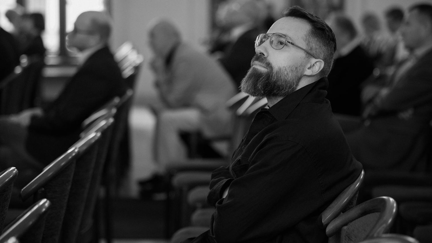 Black and white photo of a bearded man with glasses sitting in the audience at a conference.