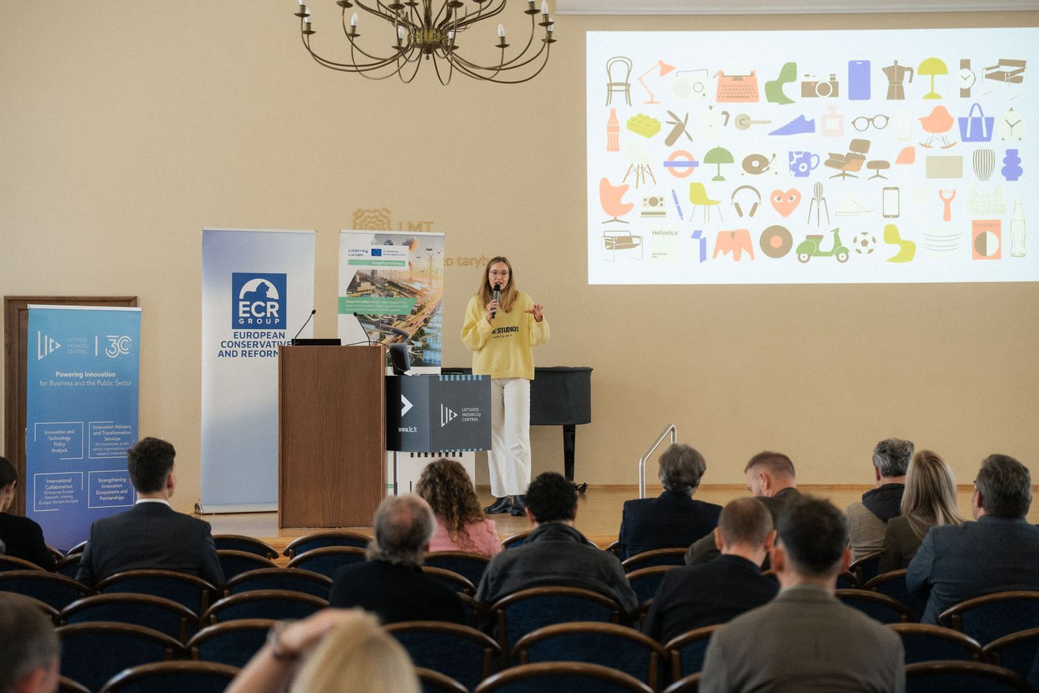 An audience watches a woman in yellow speak at a wooden podium while a screen shows colorful icons in a hall with a chandelier.