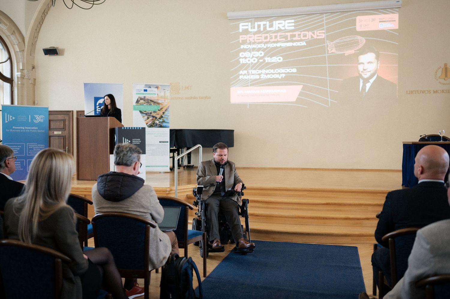Speaker at a podium and a man in a wheelchair beside a Future Predictions slide at an innovation conference.