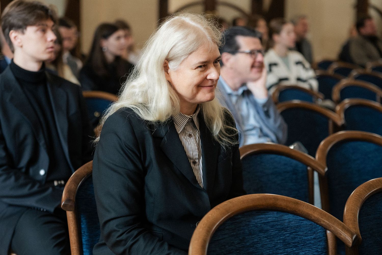 A smiling woman with long blonde hair and a black blazer sits in an audience of conference attendees in blue chairs.