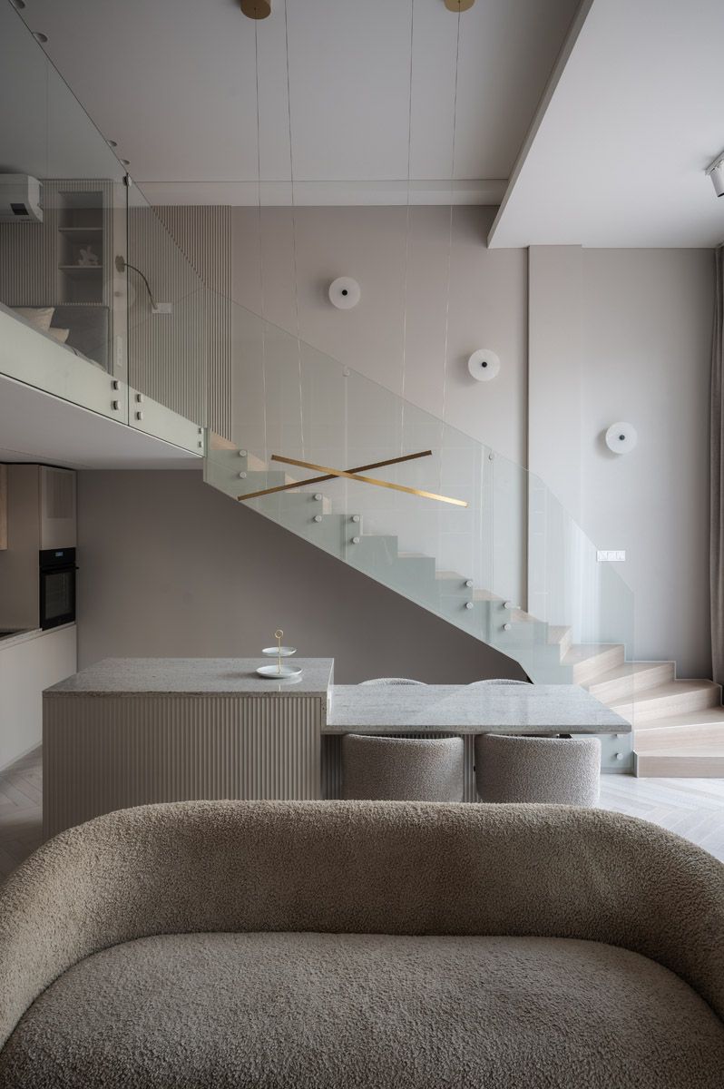 Minimalist loft interior with a glass-railed staircase, beige textured walls, marble kitchen island with stools and a boucle sofa.