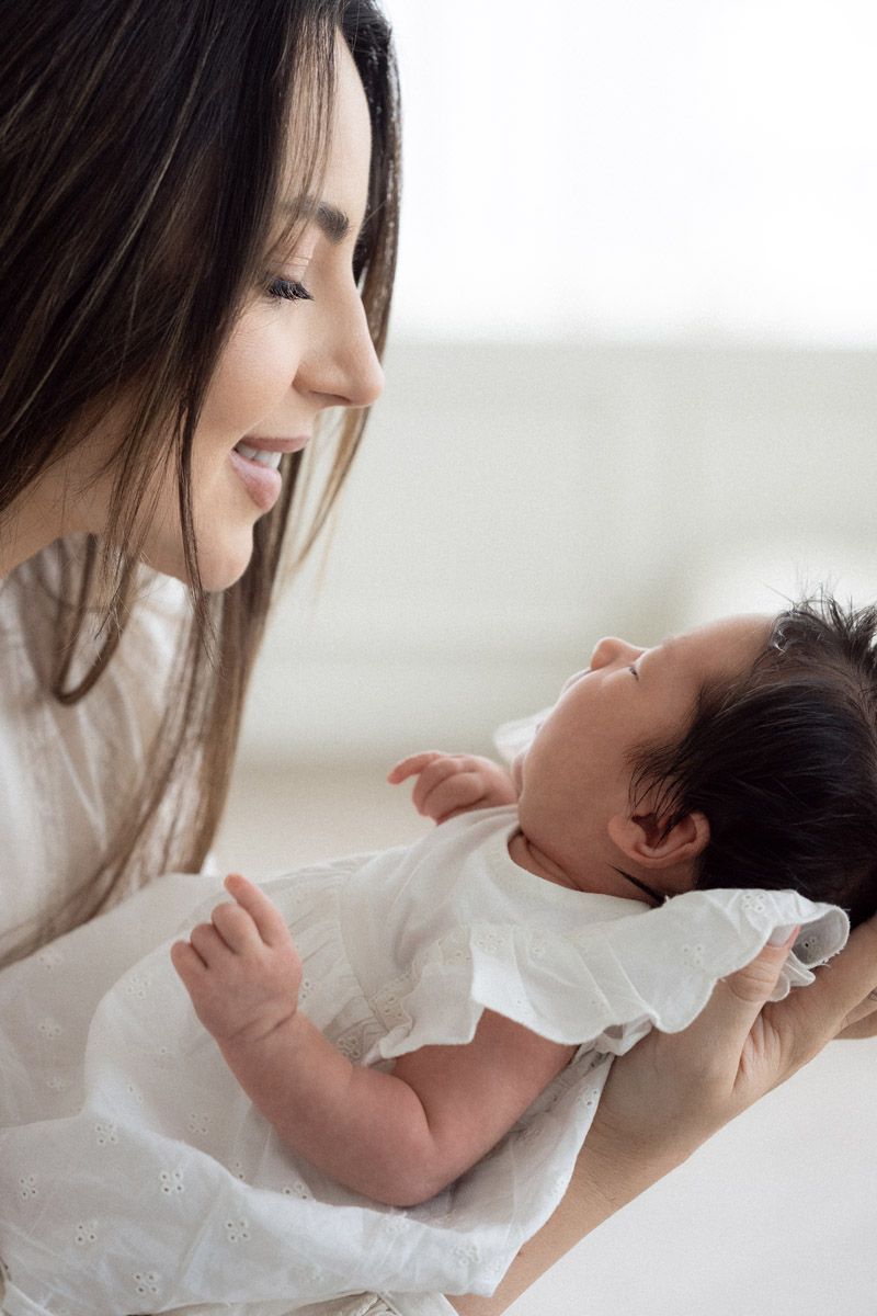 Smiling mother in white gently gazes at her sleeping newborn baby dressed in a white ruffled outfit in soft natural light.