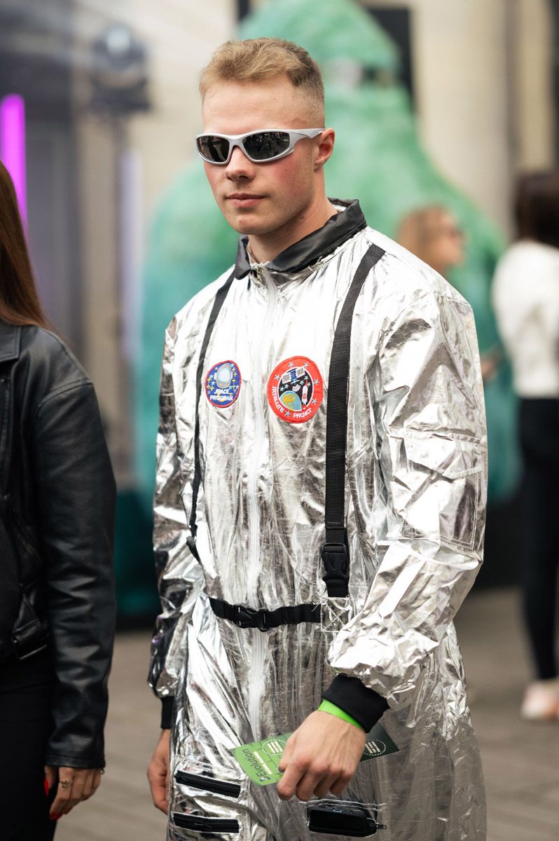 Man in a silver metallic astronaut costume with mission patches and white sunglasses stands at an outdoor event.