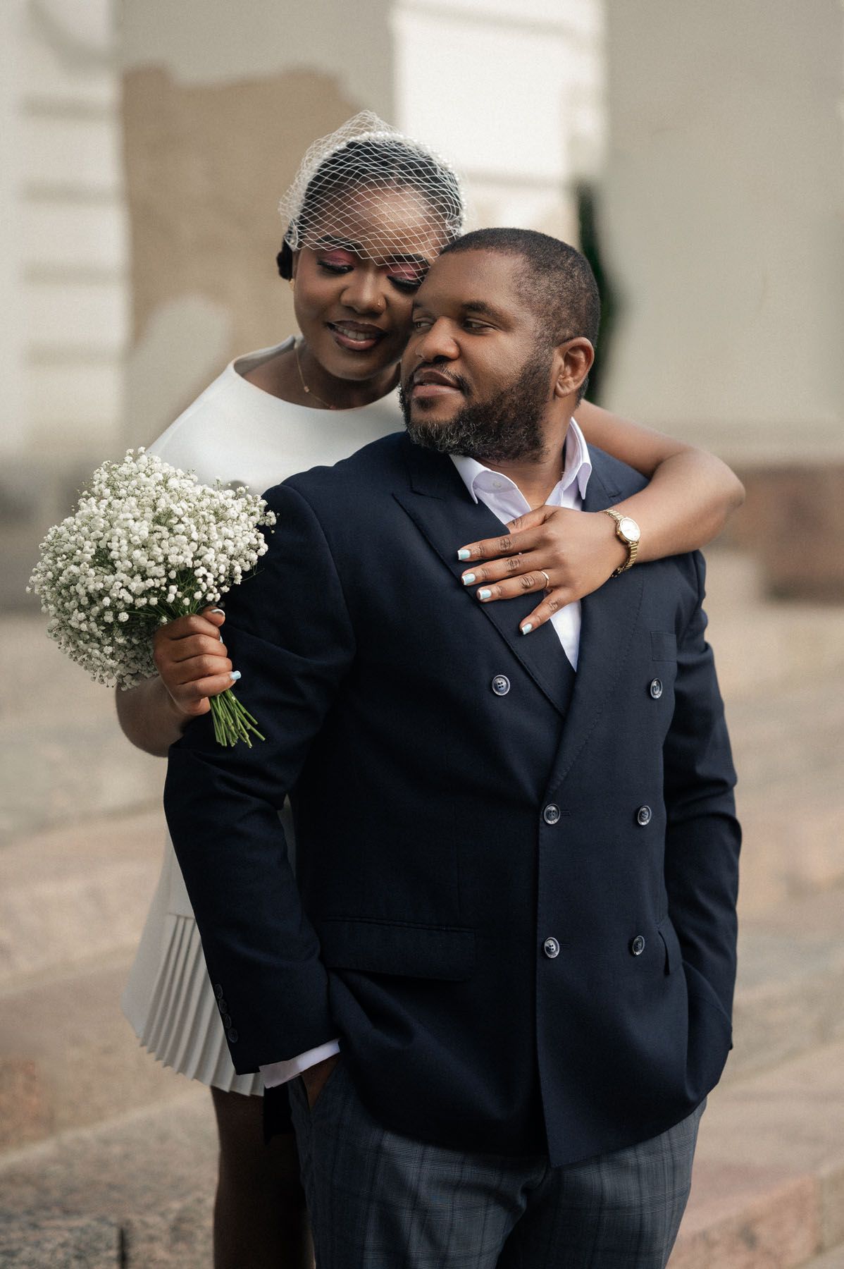 Smiling bride in a birdcage veil embraces her bearded groom in a navy double-breasted blazer, holding a baby's breath bouquet.