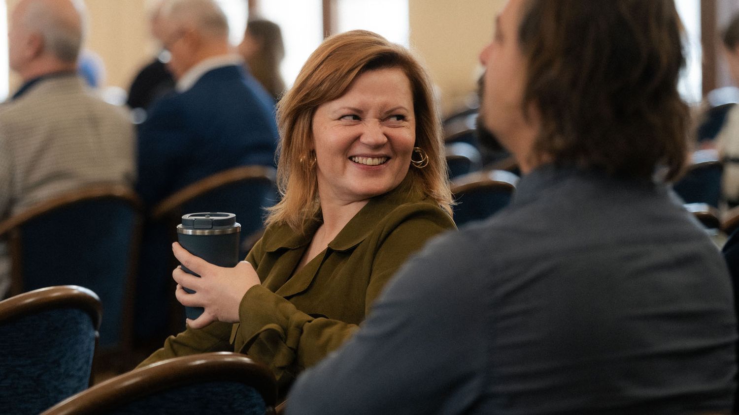 A smiling woman with red hair holds a dark cup while sitting among attendees at a conference event.