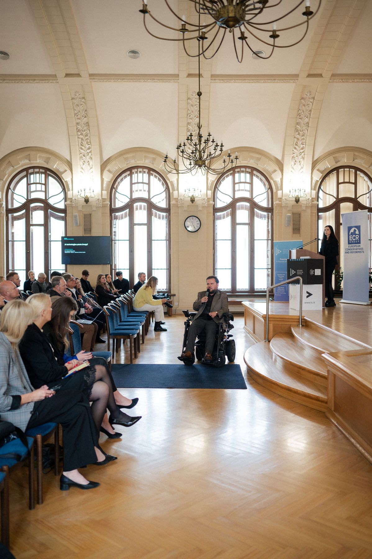 Audience seated in a grand hall with chandeliers while a man in a wheelchair and a speaker address the conference.