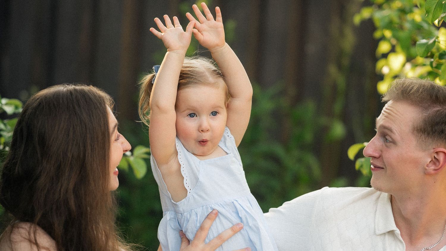 Mother holding toddler with arms raised, father smiling beside them outdoors