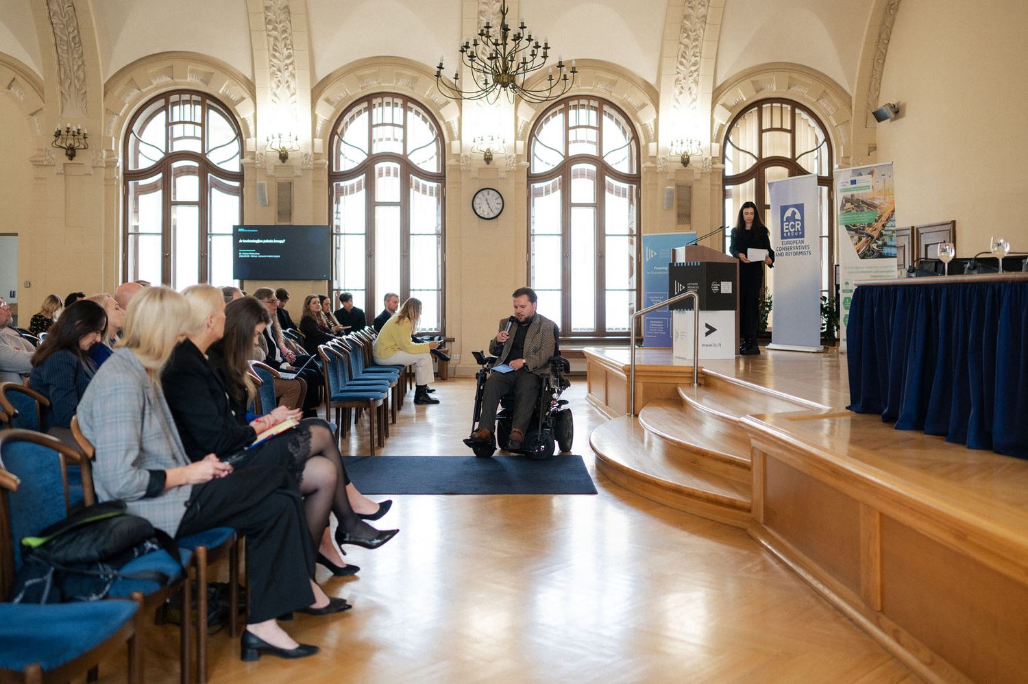 Speaker in a wheelchair addresses a seated audience on a wooden stage in a hall with tall arched windows and a podium.