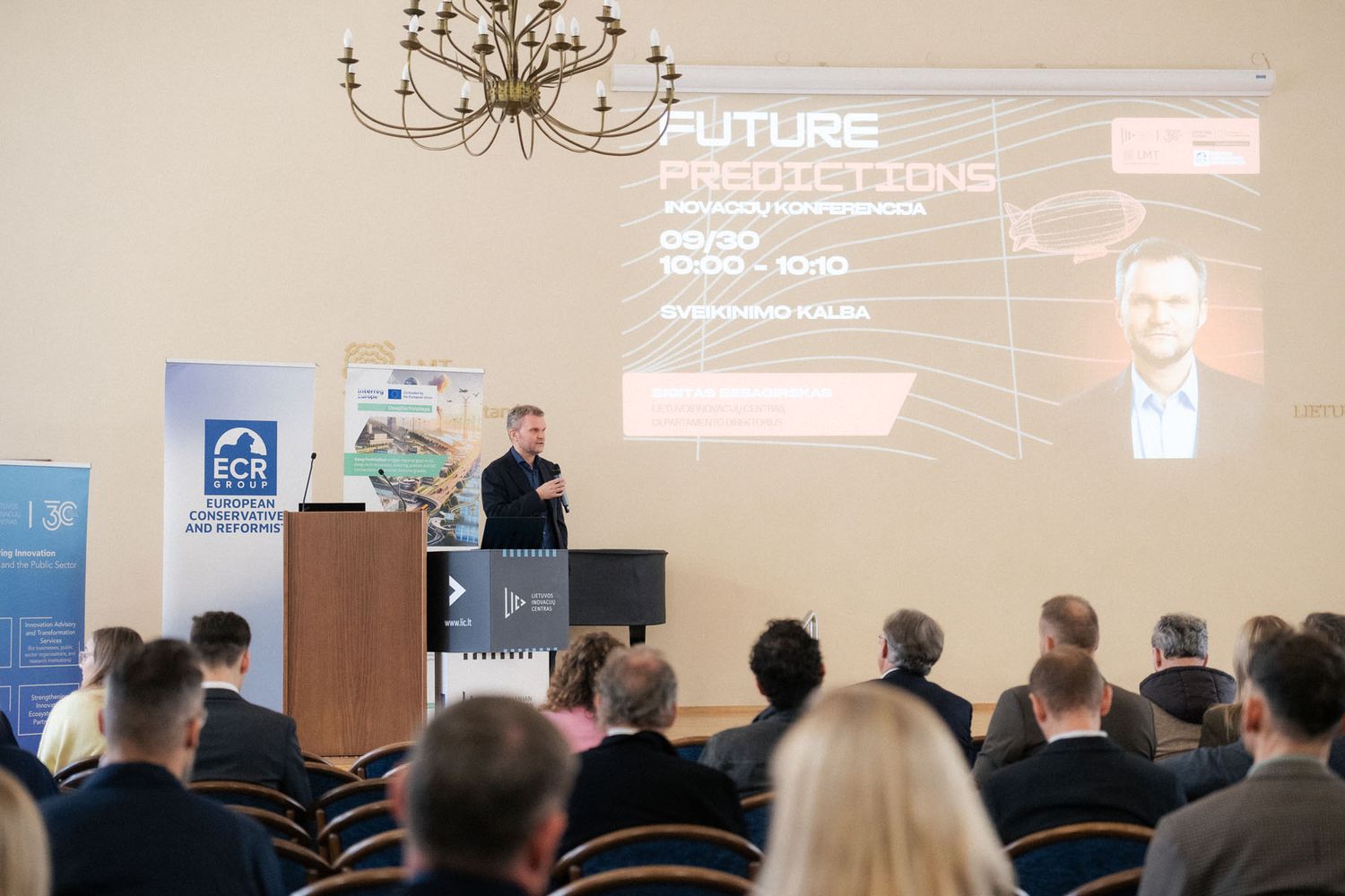 Audience members face a speaker at a podium beneath a chandelier, with a Future Predictions slide projected on the wall.