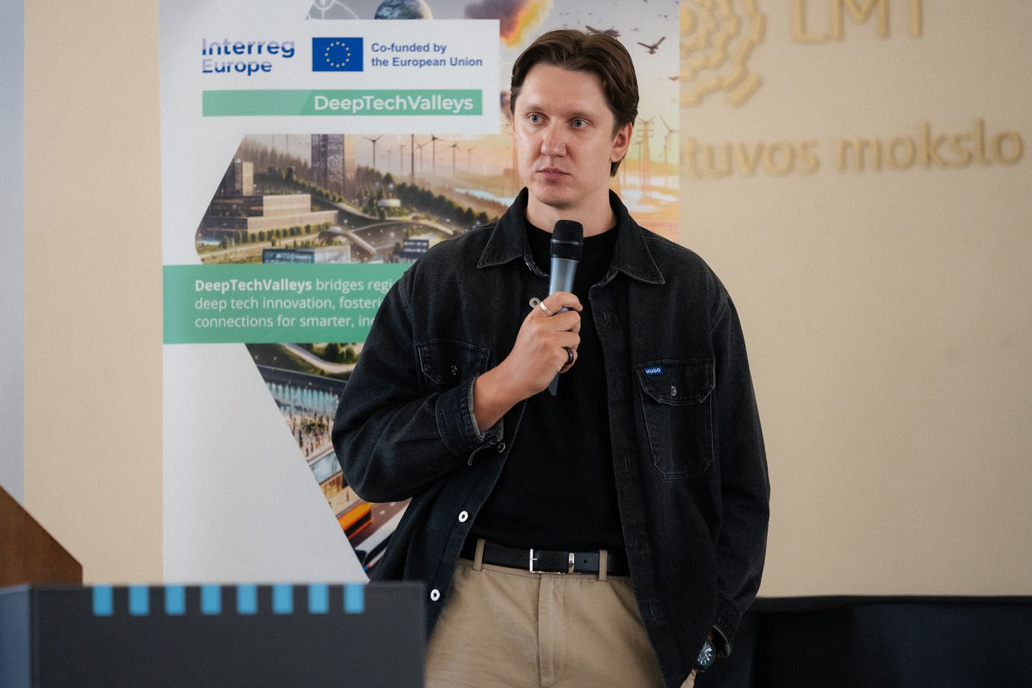 Man in a black jacket speaks into a microphone in front of a DeepTechValleys Interreg Europe banner at an innovation event.