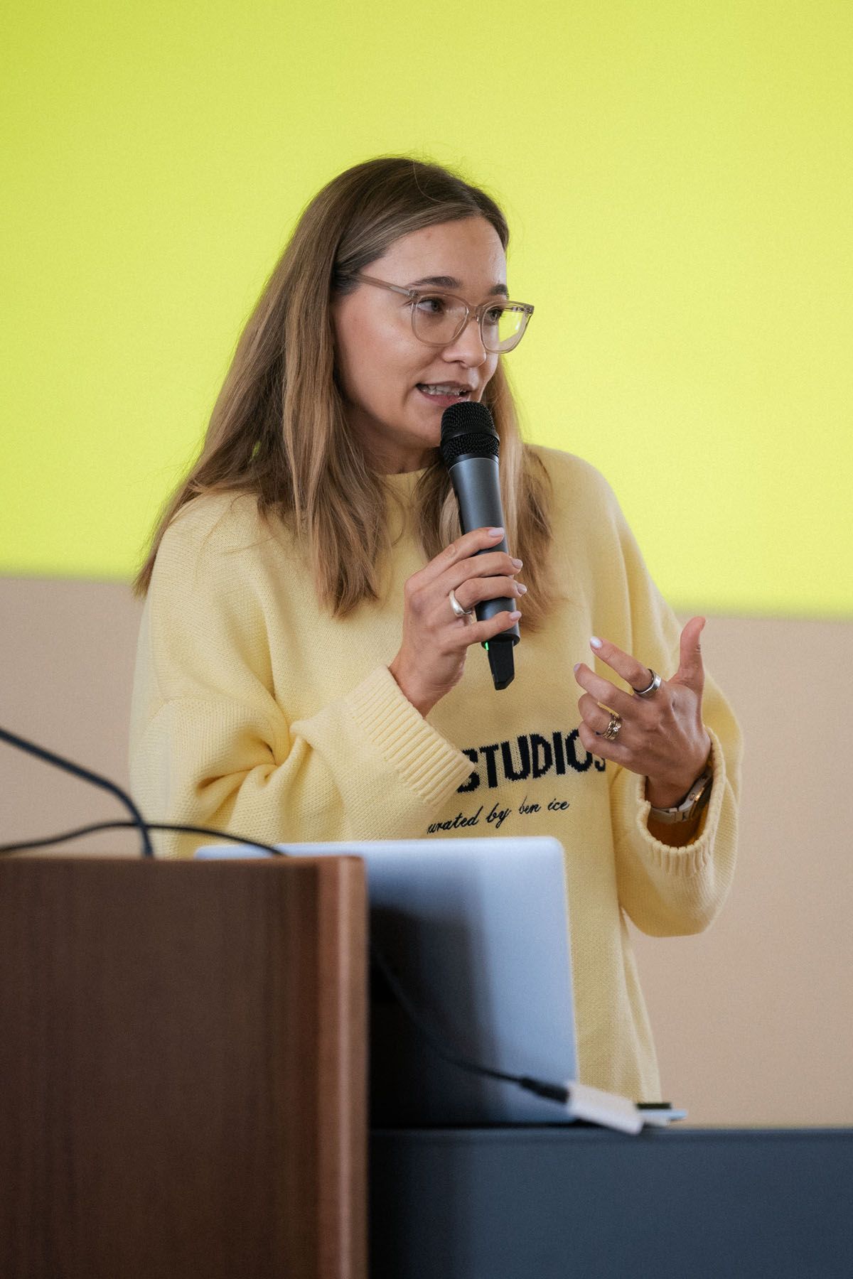 A woman with glasses and a yellow sweater gestures while speaking into a microphone at a podium during a conference.