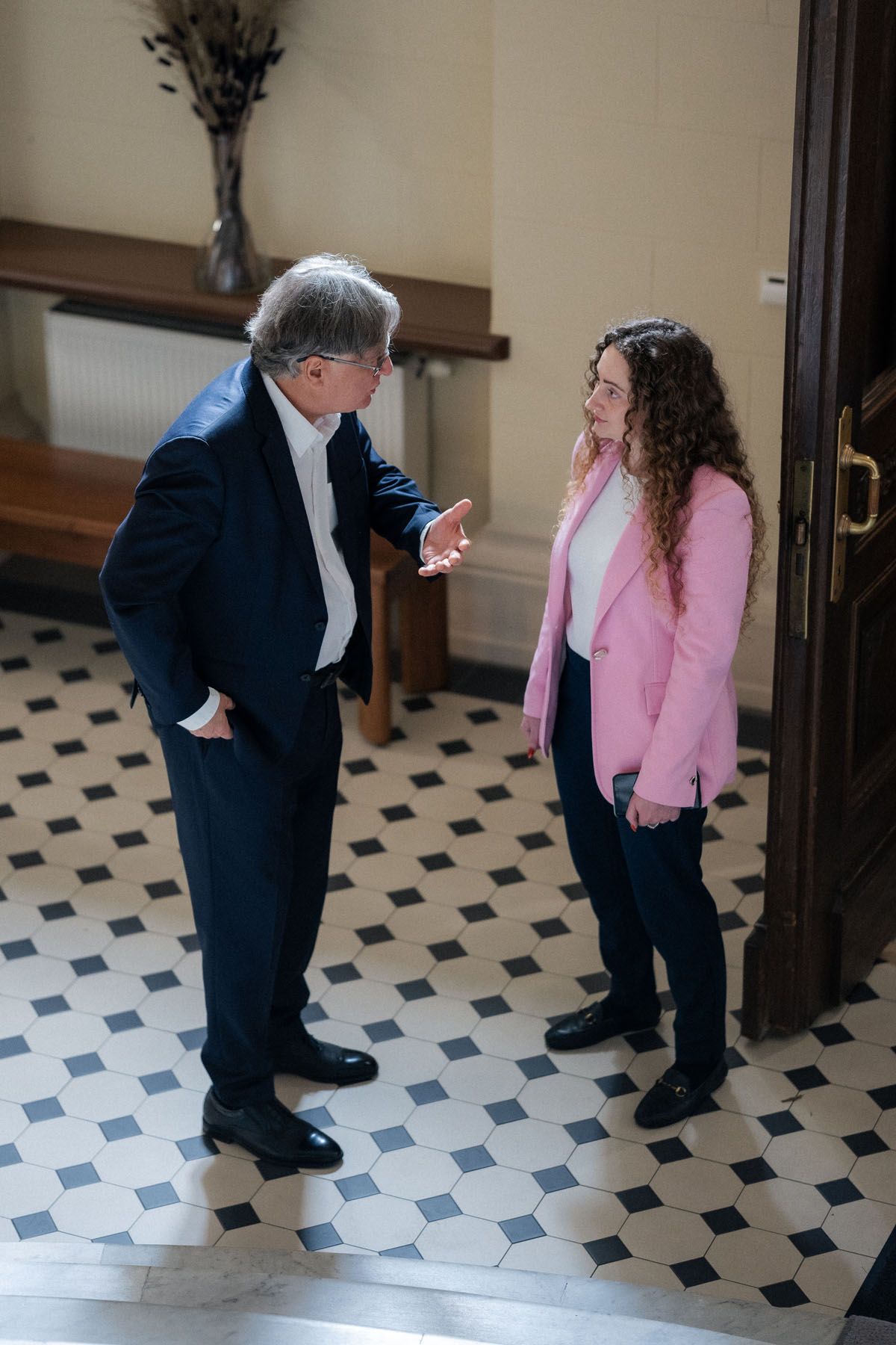 Man in a navy suit talks with a curly-haired woman in a pink blazer on a checkered tile floor at an event.