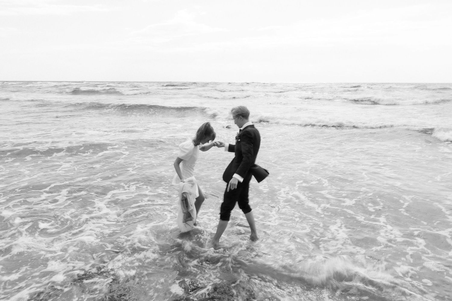 Black and white photo of a bride and groom playfully wading together in shallow ocean waves along the shoreline.