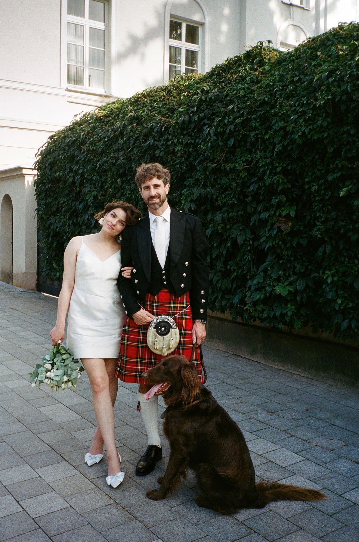 Bride in a white dress with a bouquet stands beside a groom in a red tartan kilt and a brown dog on a paved street.