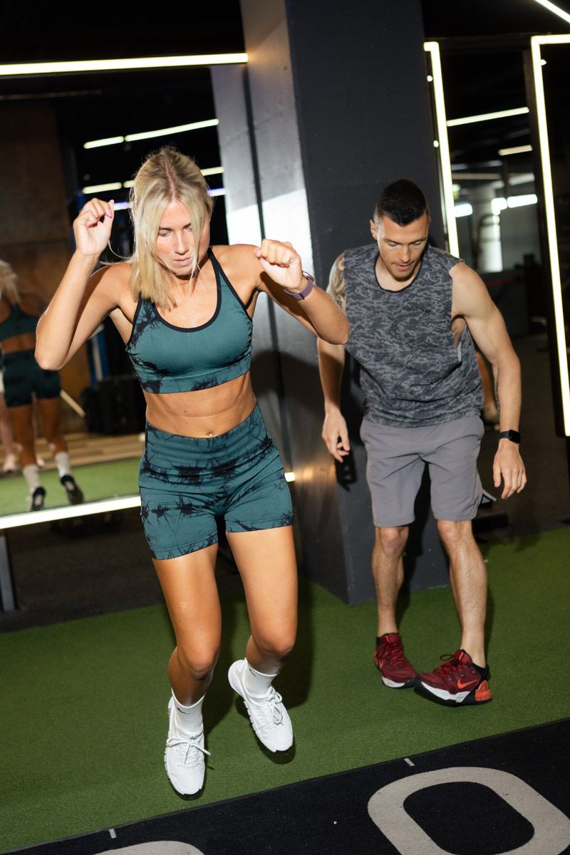 Woman in a teal sports bra and shorts jumps during a gym workout while a male trainer in a gray tank top watches behind her.