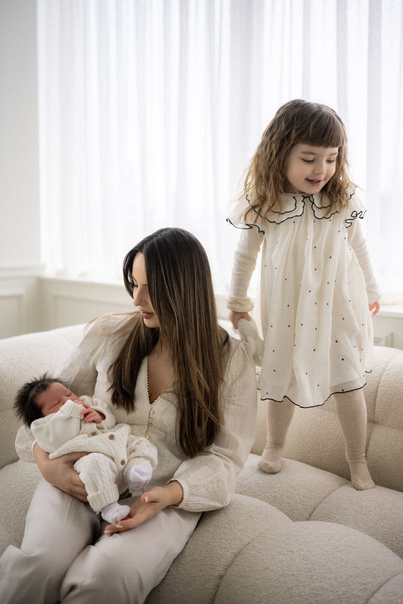 Mother holding newborn as daughter in knit cardigan stands nearby, family studio session