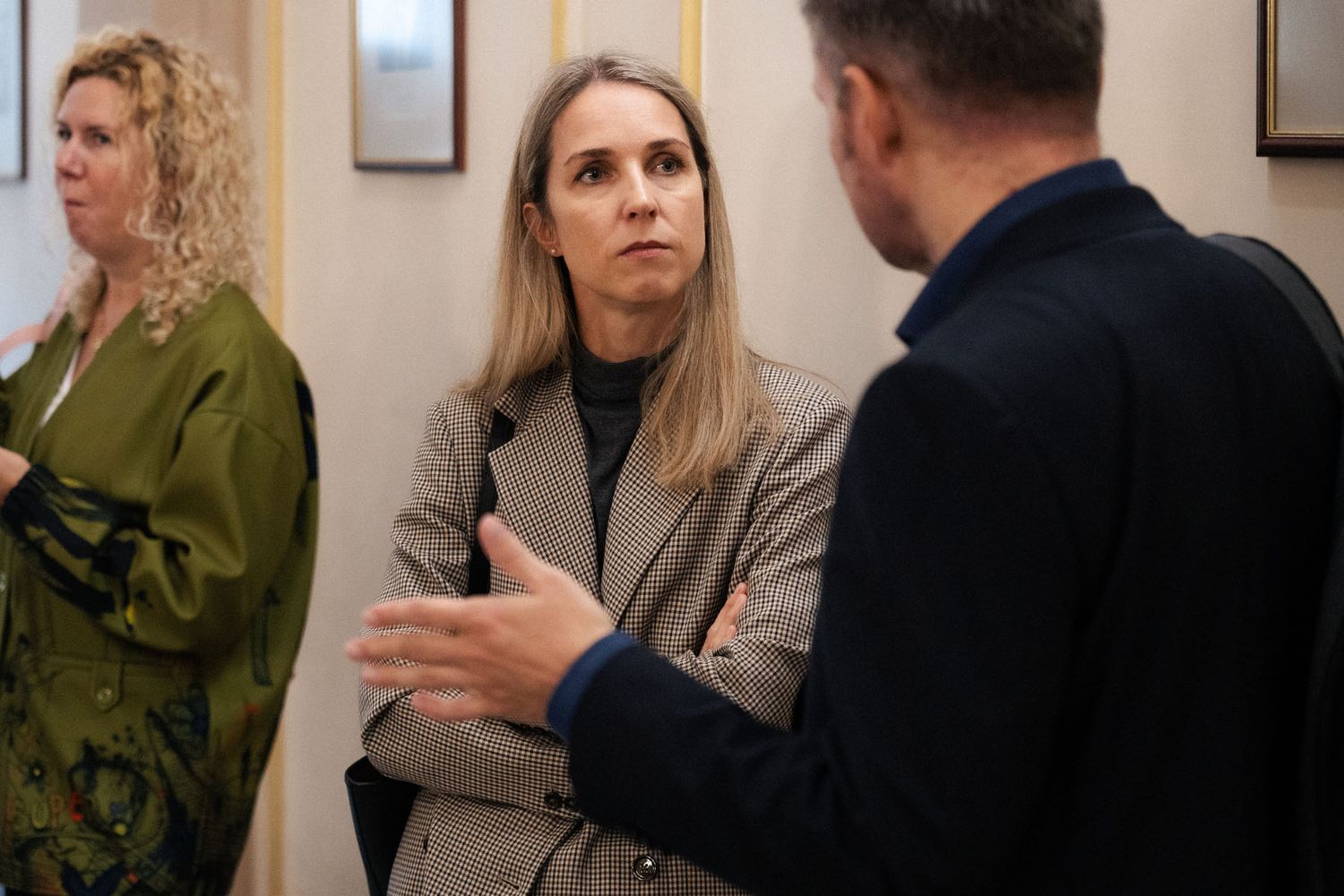 A blonde woman in a tweed blazer listens attentively while a man in a dark jacket gestures during a conversation indoors.