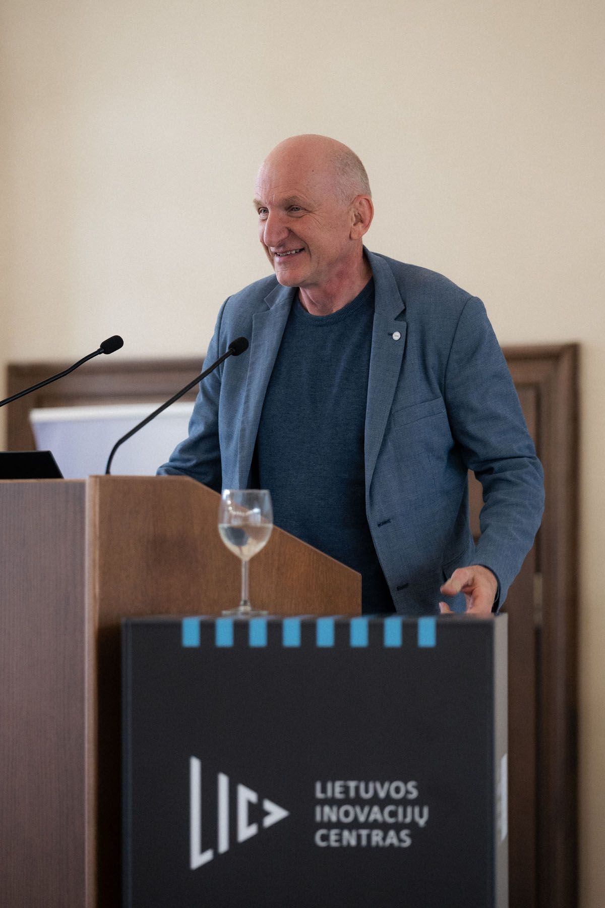 A smiling bald man in a blue blazer speaks at a wooden lectern with the Lietuvos inovacijų centras logo and a glass of water.