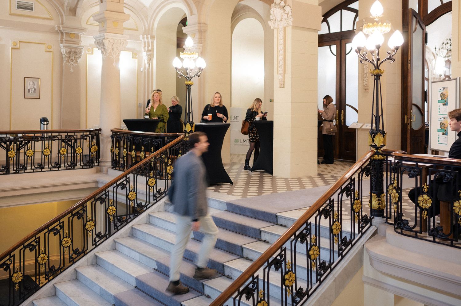 Visitors and staff gather on a marble staircase with ornate gold-detailed railings in a historic building lobby.