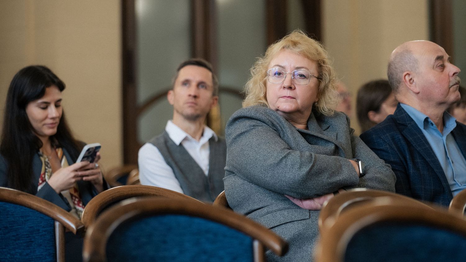 Attendees sit in blue chairs at a conference, including a woman with glasses and a woman looking at her phone.