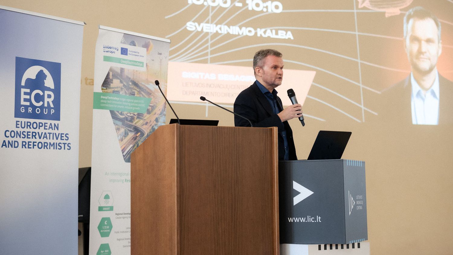 A speaker addresses the audience from a wooden podium at an ECR Group conference, with a projected presentation slide behind him.