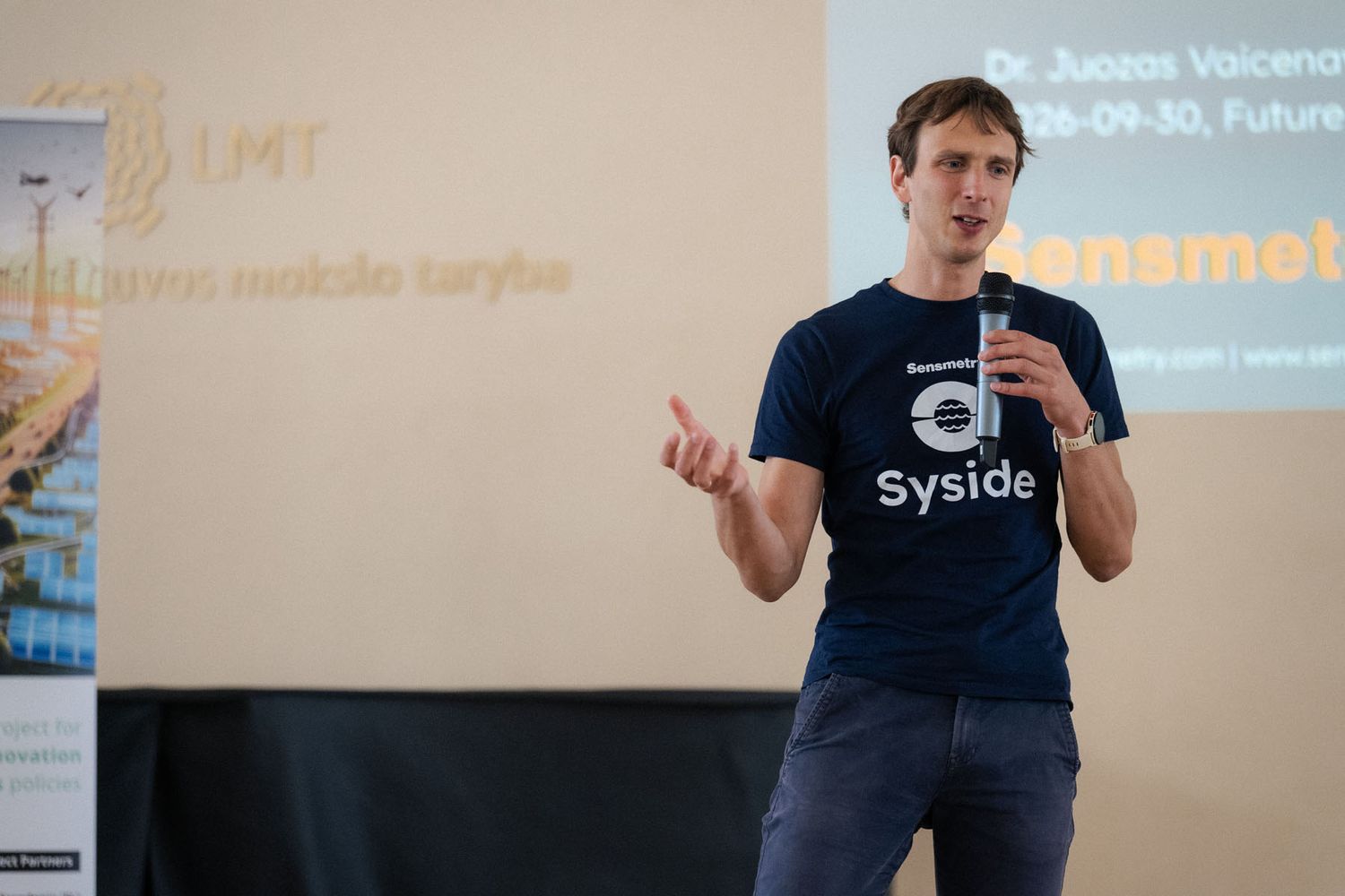 Smiling speaker in a Syside t-shirt gestures with a microphone while presenting in front of a projected slide.