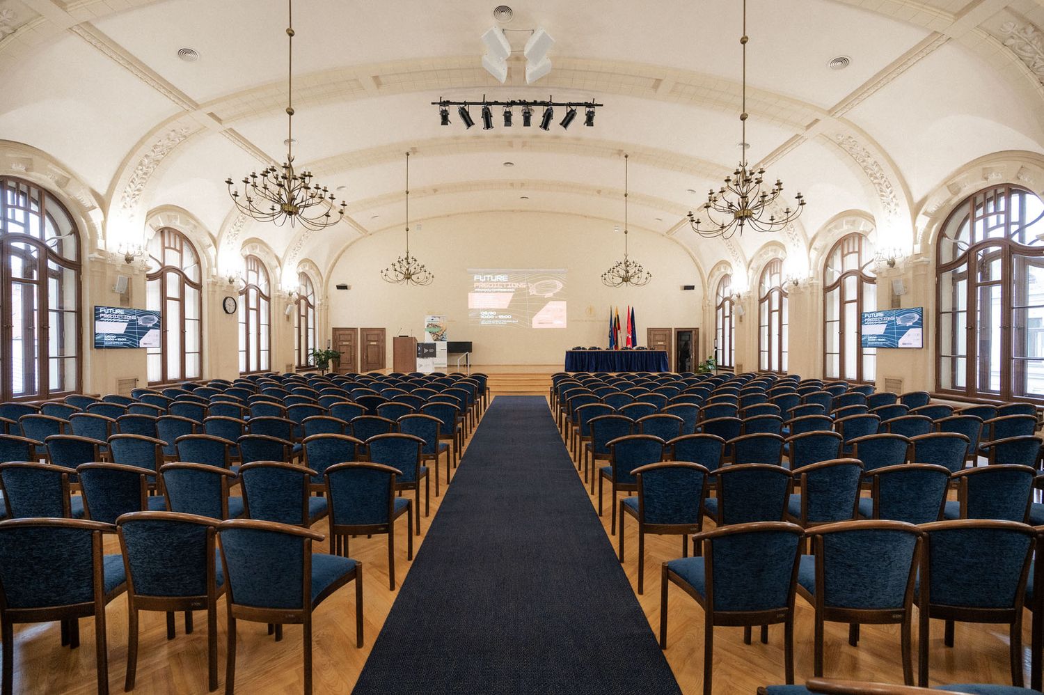 Rows of blue chairs line a central aisle in an ornate conference hall with chandeliers and arched windows.