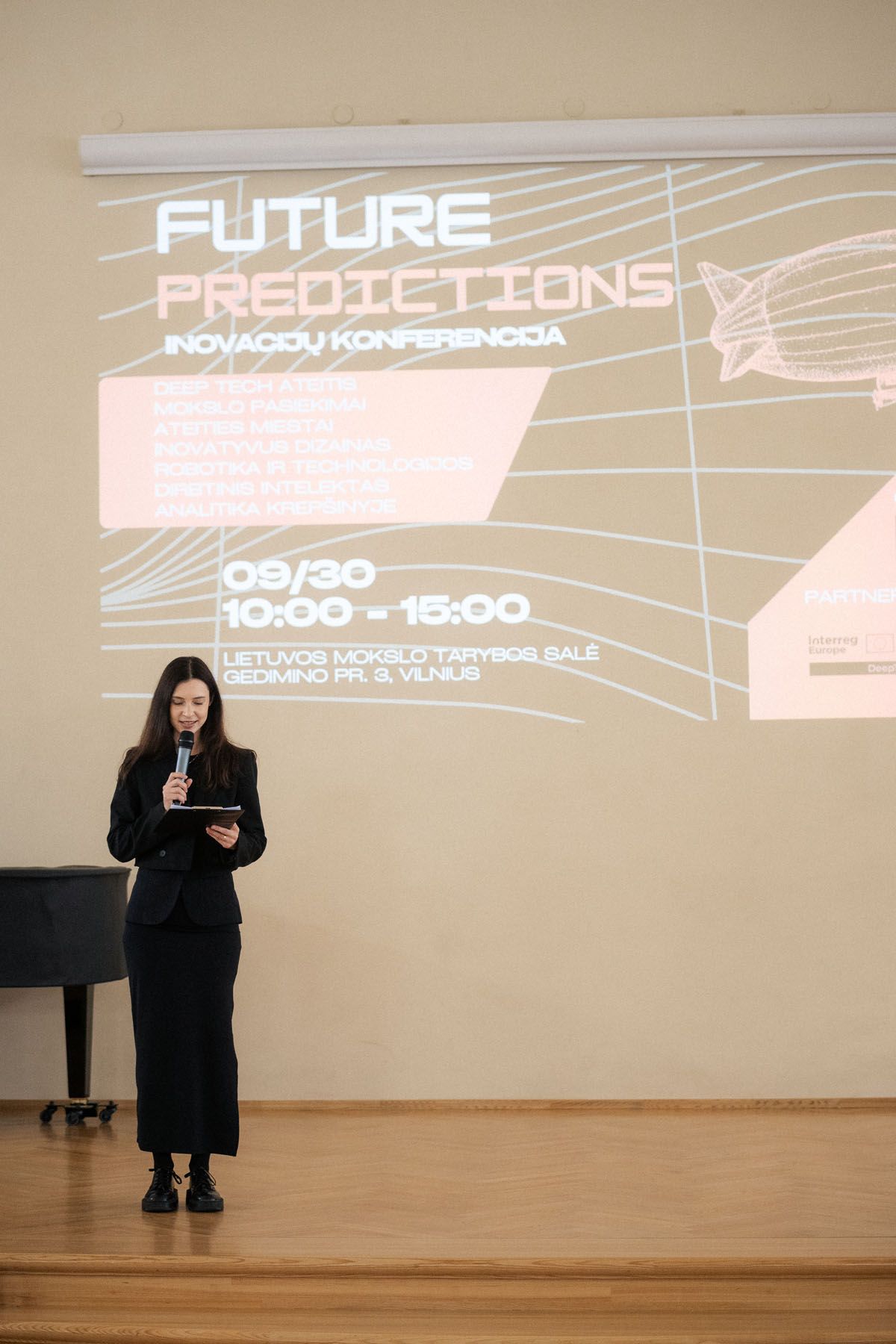 A woman in black speaks into a microphone on stage in front of a Future Predictions innovation conference projection in Vilnius.