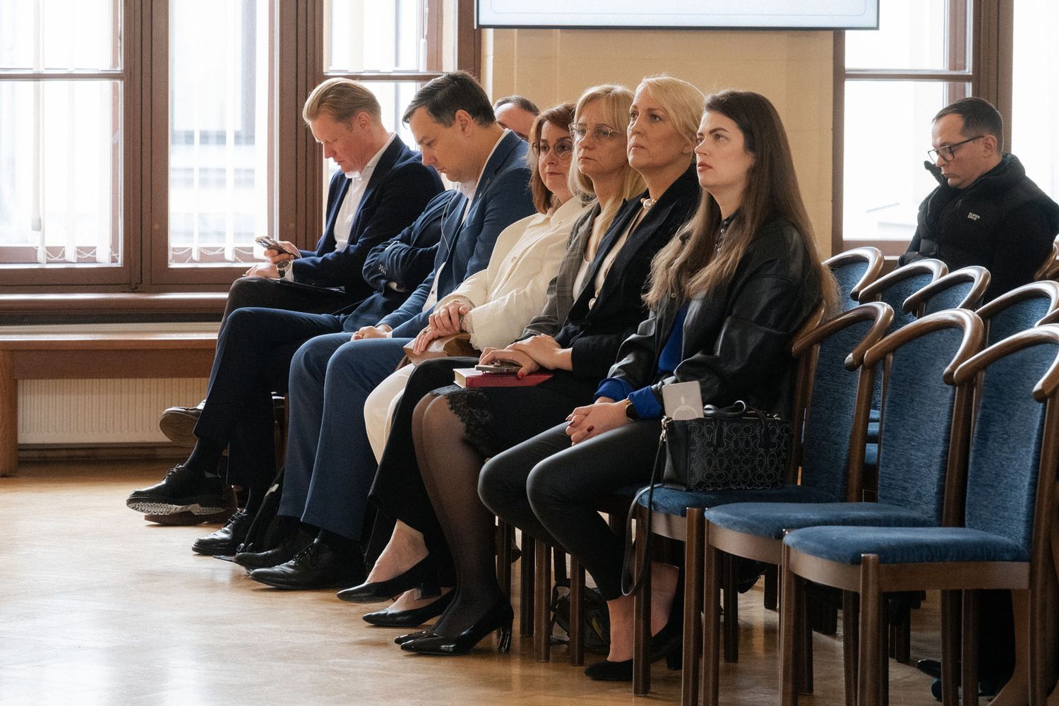 Row of attendees in business attire sit on blue chairs beside tall windows, waiting for a conference session.