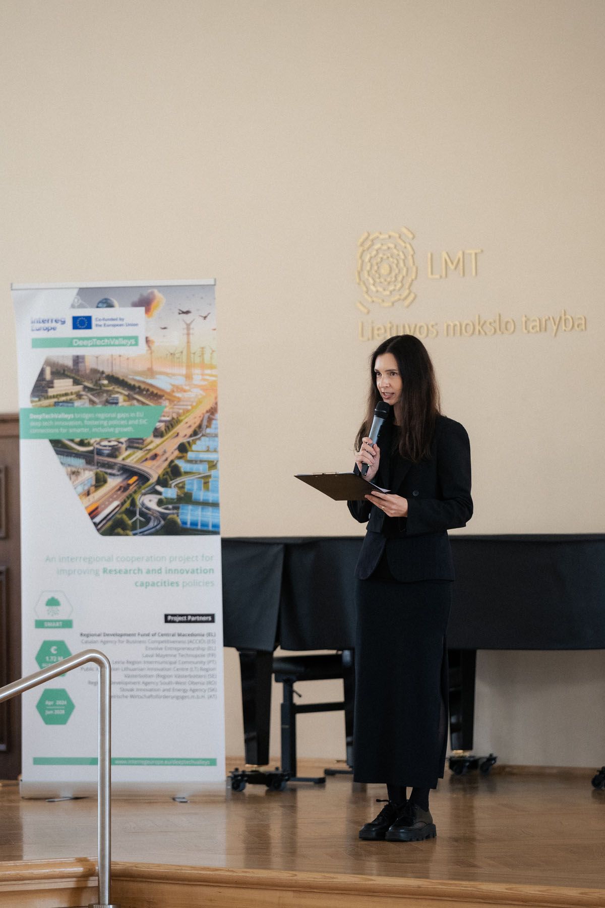 A woman in black holds a microphone and folder while speaking on stage beside an EU-funded research and innovation banner.
