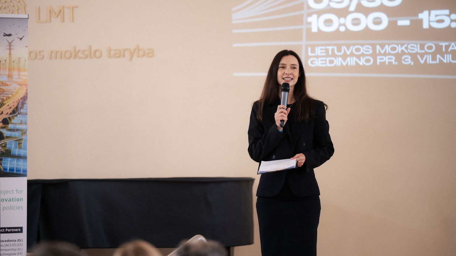 A woman in a black suit holds a microphone and speaks on stage in front of a projected screen at a Vilnius science event.