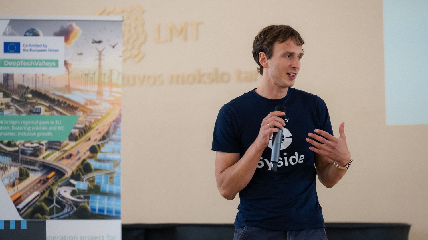 A young man in a Syside t-shirt holds a microphone and gestures while presenting near a DeepTechBridge banner.