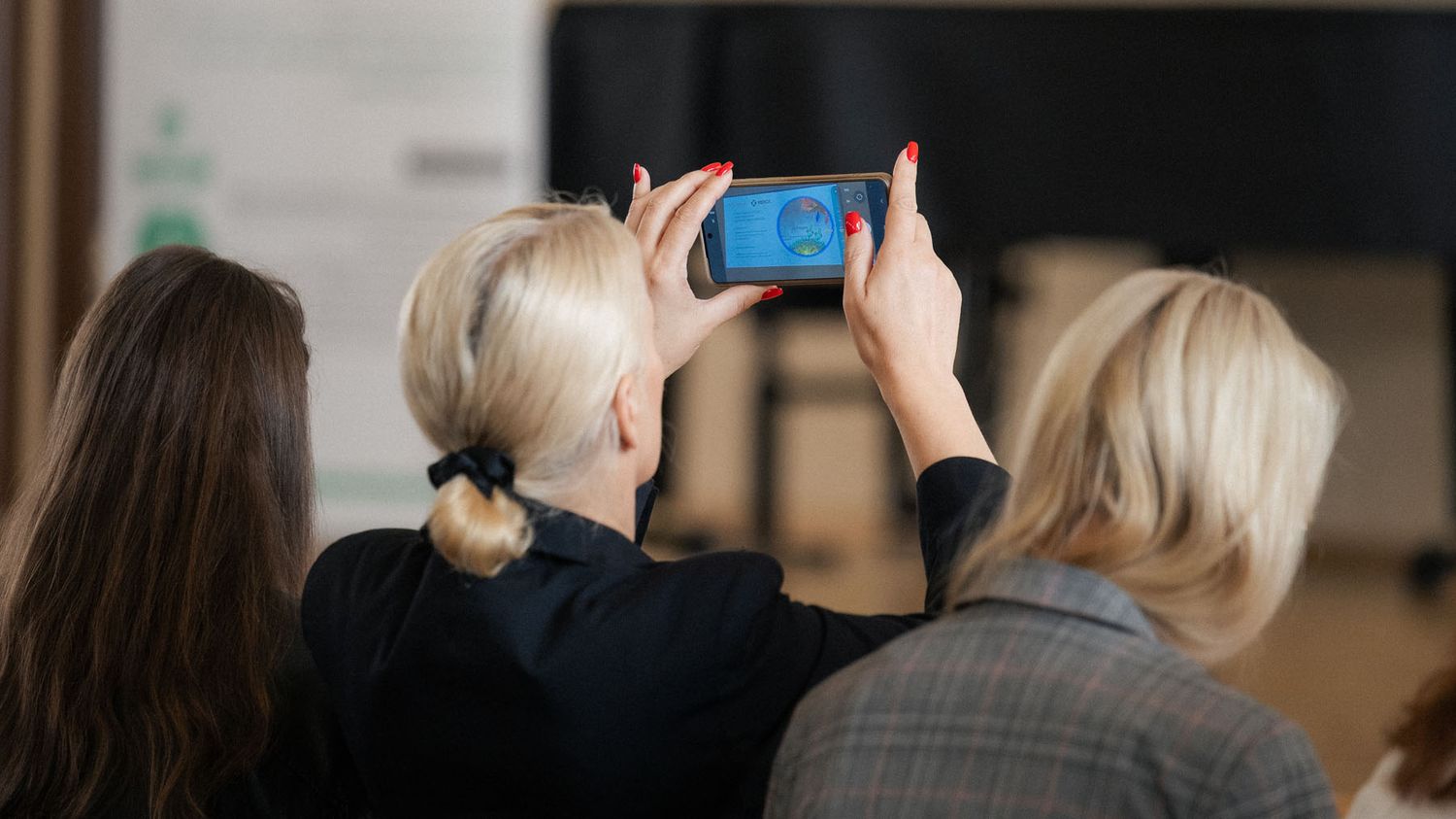 Woman in a dark blazer photographs a stage with her smartphone while two other attendees stand beside her.