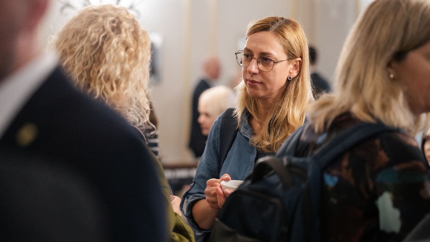 A woman with glasses and shoulder-length blonde hair stands among attendees at a busy indoor conference event.