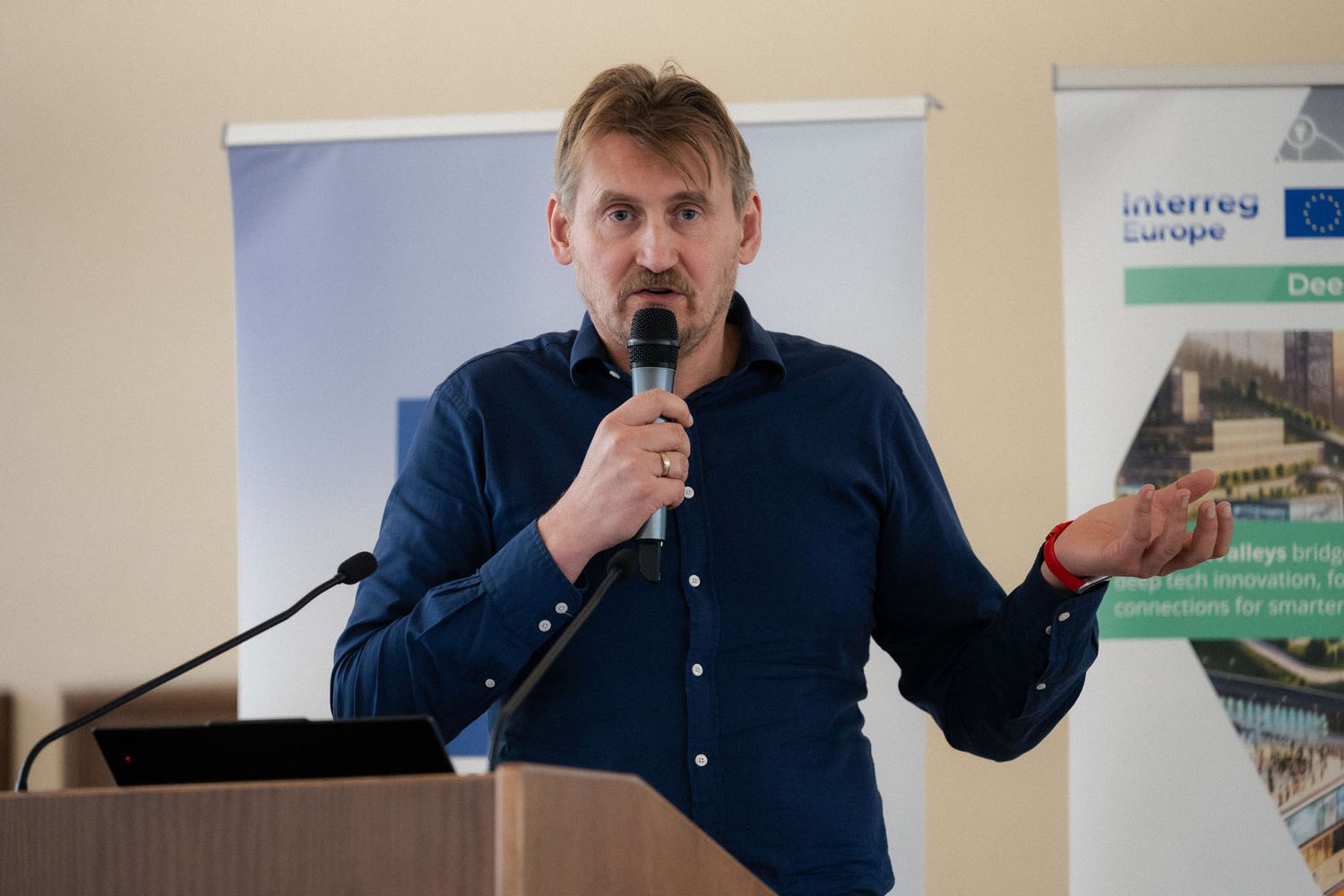A man in a navy shirt holds a microphone and gestures with one hand while speaking at a conference podium.