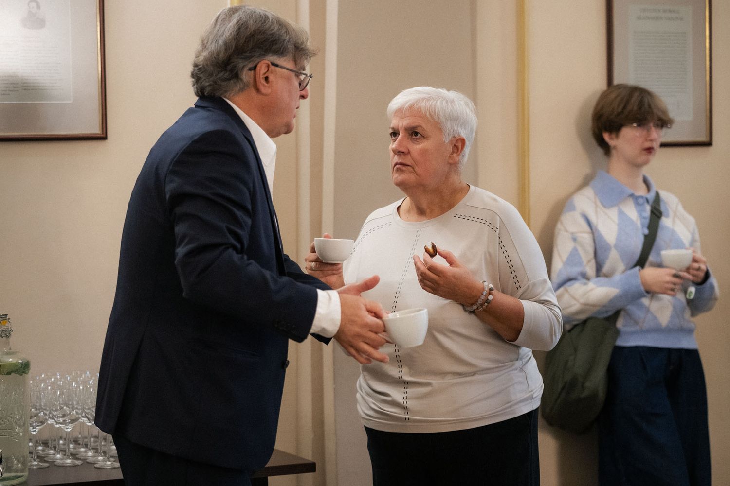 A man in a navy blazer and a gray-haired woman in a cream sweater chat over coffee cups while a young person looks on.